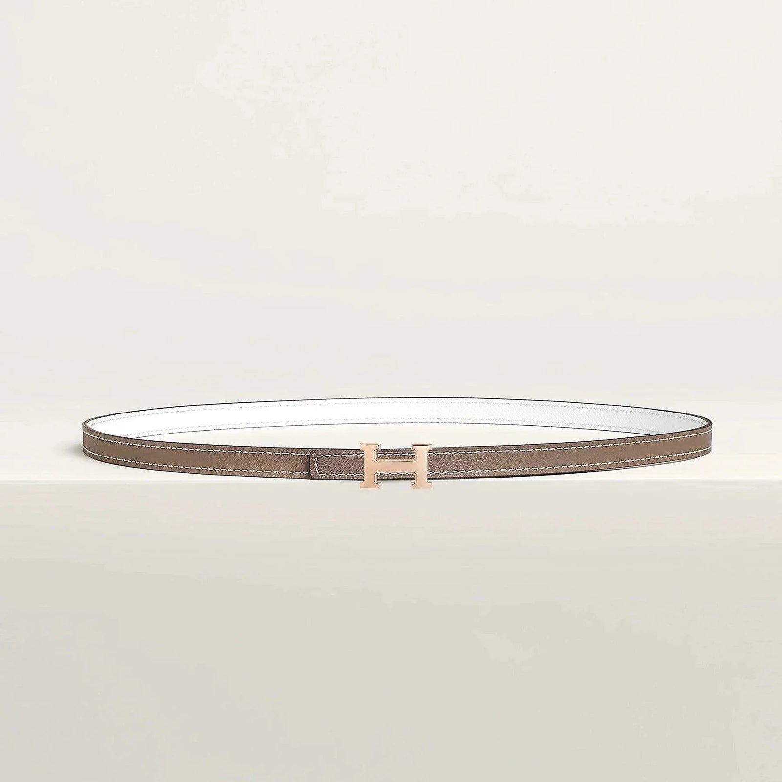 Hermes belt with elegant gold buckle on beige background, showcasing luxury and minimalist design.